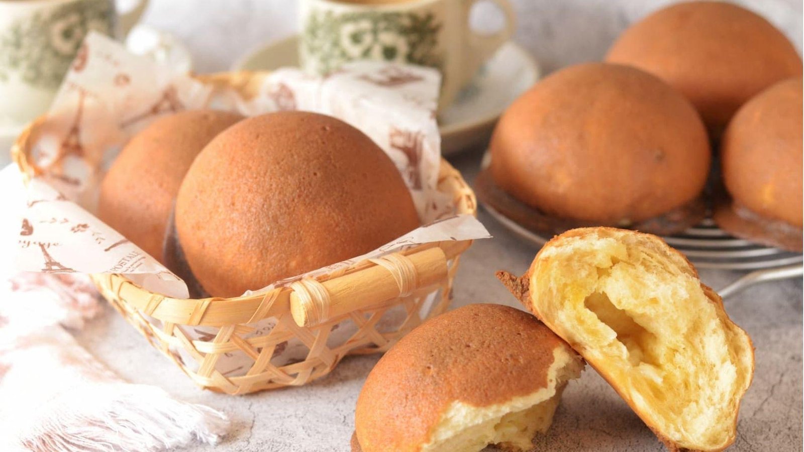 Golden coffee buns in a wicker basket with one split open revealing a soft buttery interior, beside two cups of coffee.