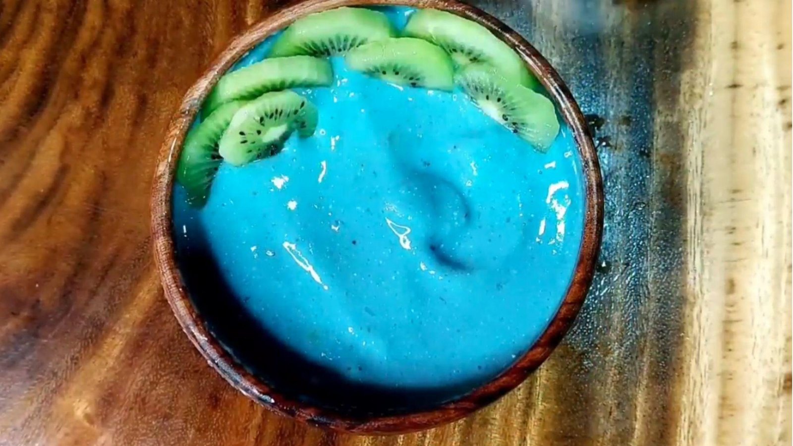Overhead view of a vivid blue spirulina smoothie bowl in a wooden bowl topped with sliced kiwi along one edge.