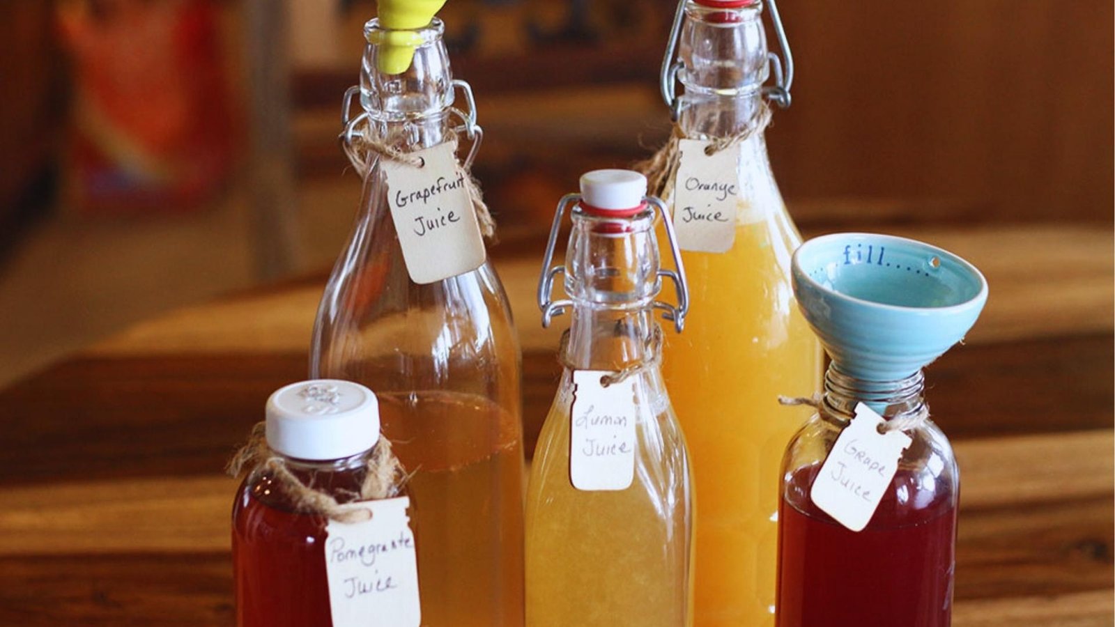 Five flip-top glass bottles filled with colorful homemade kombucha labeled grapefruit, orange, lemon, grape, and pomegranate juice on a wood table.