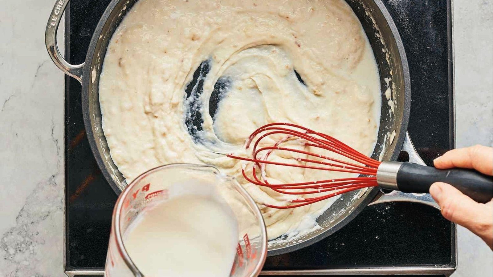 Hand whisking thick cream sauce while pouring milk into pan, step for pasta cream sauce base
