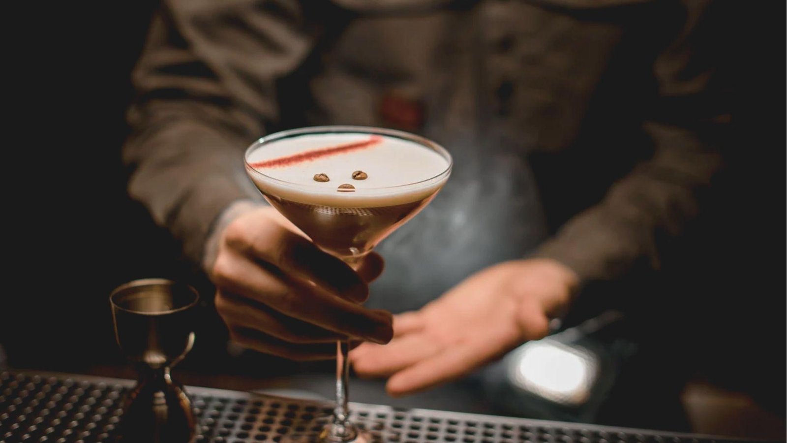 Bartender presenting a frothy Mezcal Espresso Martini in a classic martini glass, topped with three coffee beans and a dusting of spice.