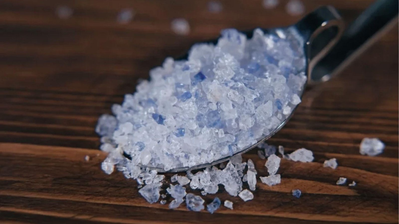 Coarse blue salt crystals for blue salt trick recipe in metal scoop on dark wood with salt scattered