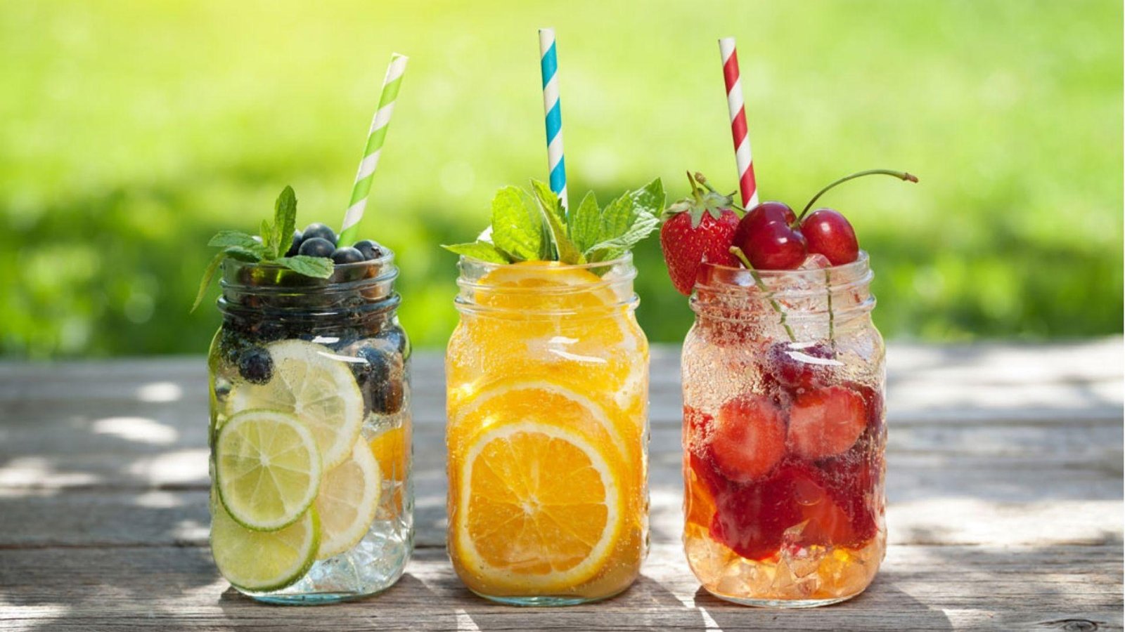 Three mason jars of fruit-infused water with blueberry-lime, orange-mint, and strawberry-cherry, each with colorful striped straws outdoors.