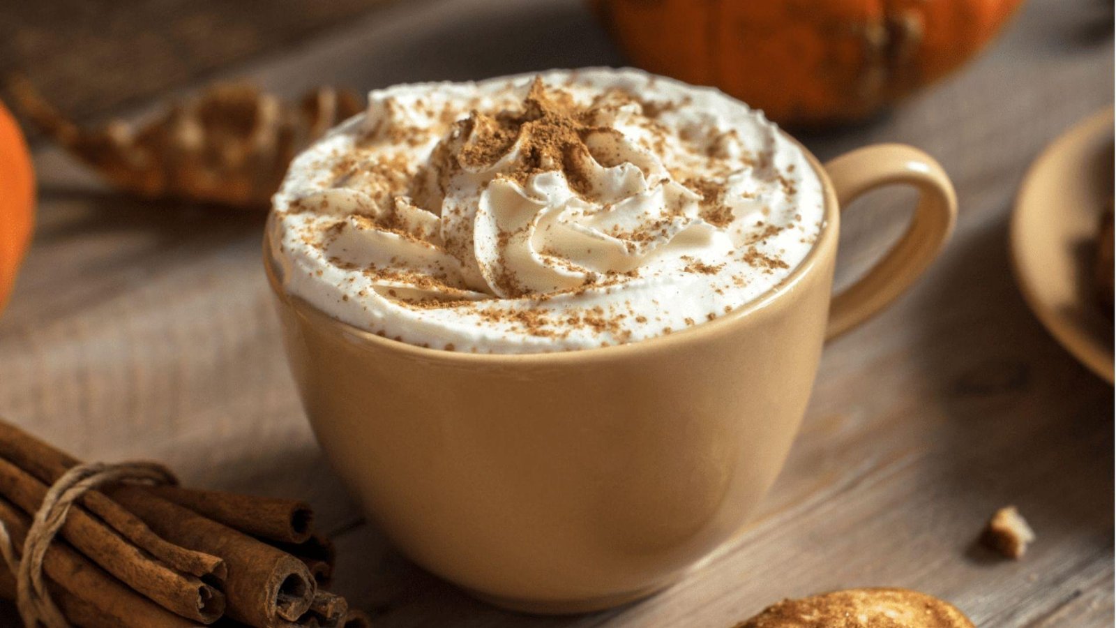 A tan ceramic mug of pumpkin spice latte topped with whipped cream and cinnamon, with pumpkins and cinnamon sticks nearby.