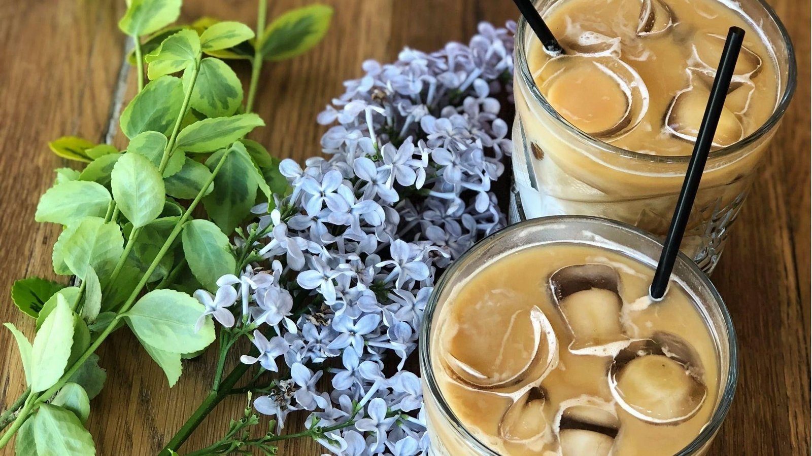 Two iced latte glasses with black straws on a wooden table surrounded by blooming purple lilac flowers.