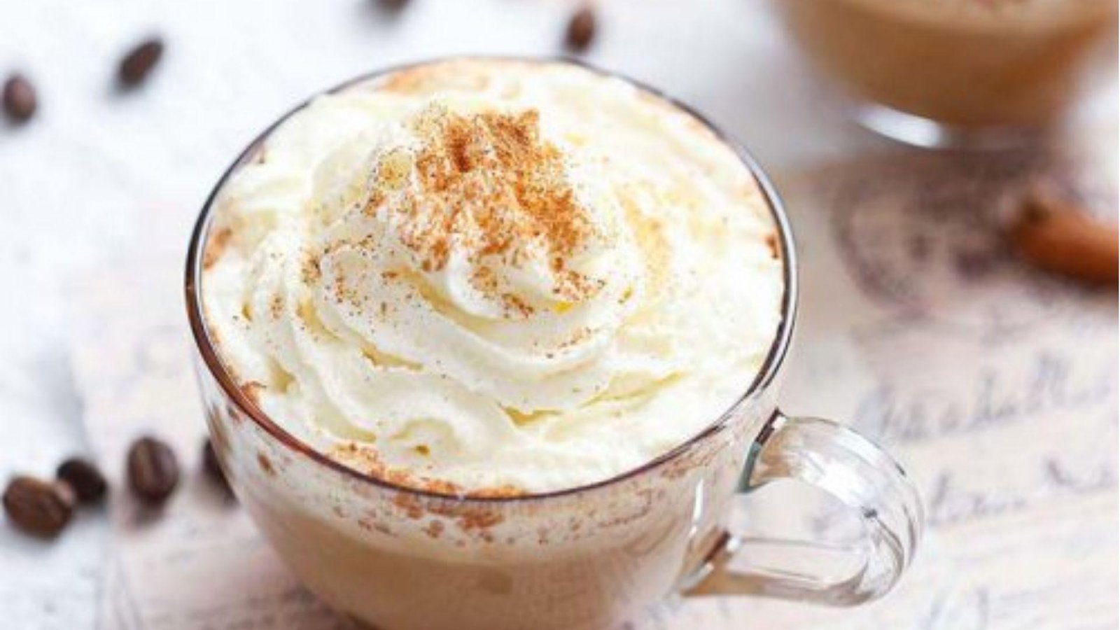 A glass mug of creamy coffee topped with a swirl of whipped cream dusted with cinnamon, with coffee beans nearby.