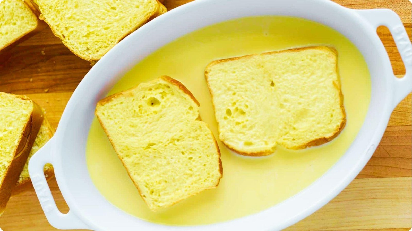 Two slices of wheat bread soaking in a yellow egg and milk custard mixture in a white dish.