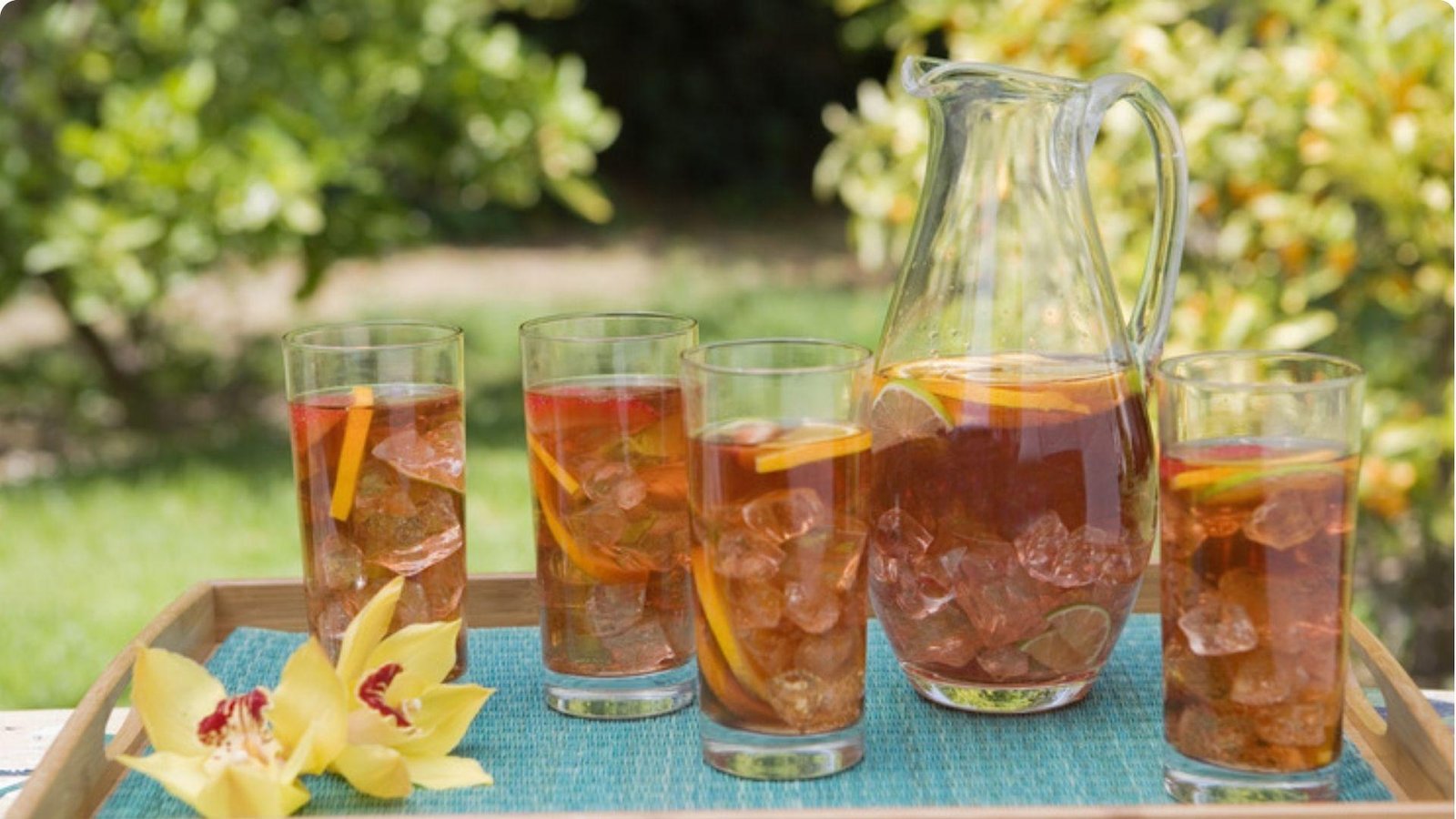Four glasses of fruity iced tea with a glass pitcher on a tray outdoors, garnished with citrus and a yellow orchid.
