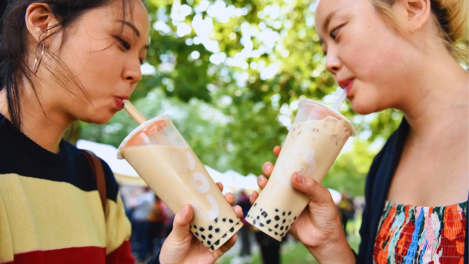 Two women sip iced milk tea with black tapioca pearls through straws outdoors, clinking plastic cups under sunlit green trees.
