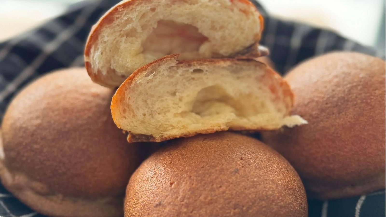 A stack of golden-brown coffee buns on a checkered cloth with one bun split open showing a fluffy butter-filled interior.