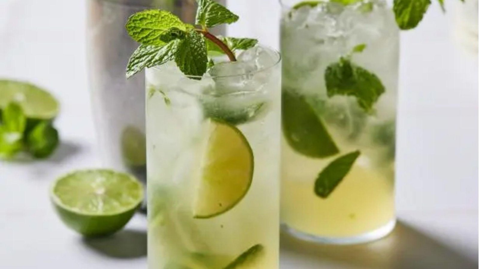 Two tall glasses of clear mojito with lime slices, fresh mint, and crushed ice on a white surface with lime halves.