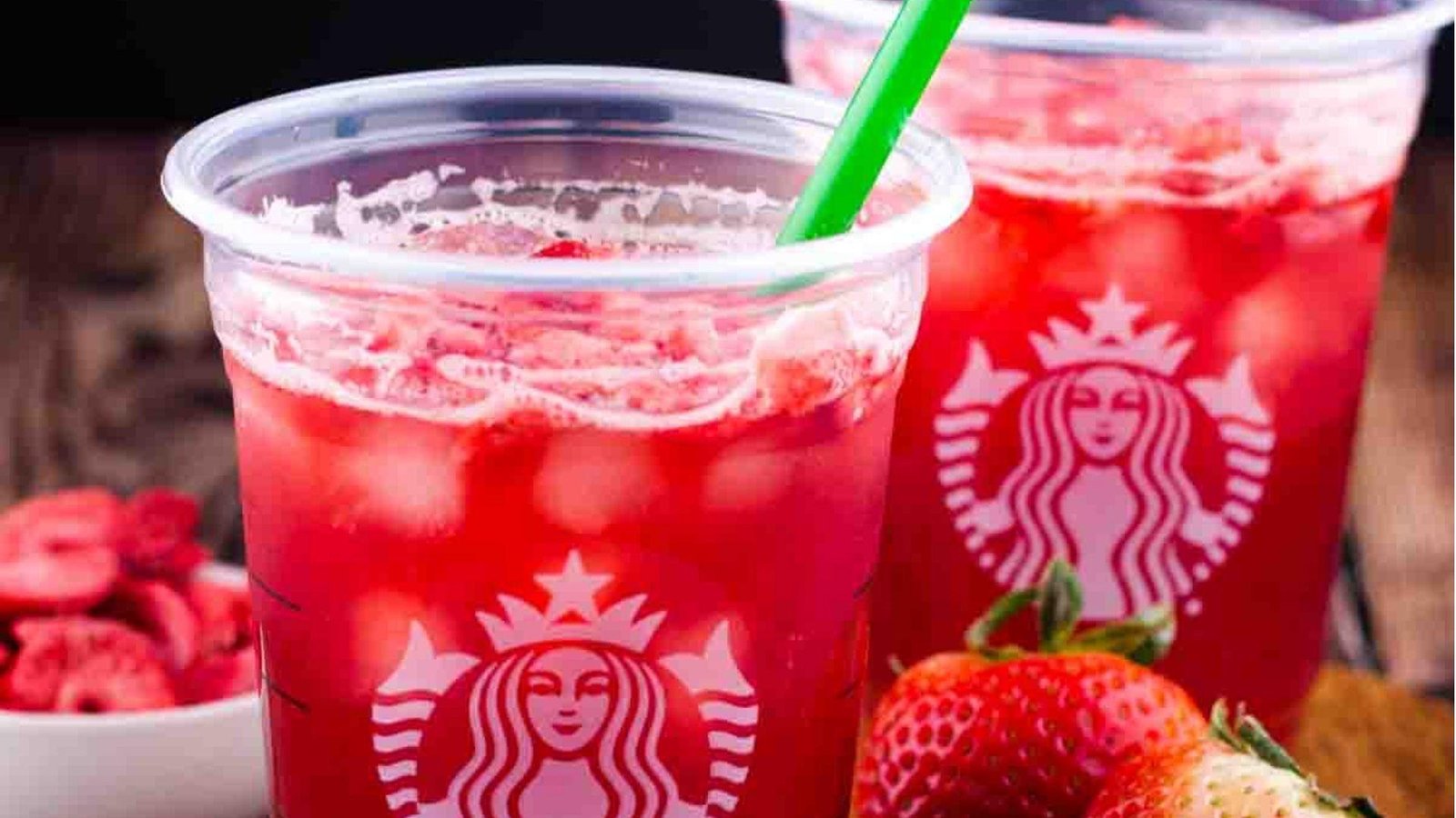 Two Starbucks cups filled with vibrant red strawberry iced drinks with a green straw, surrounded by fresh strawberries.