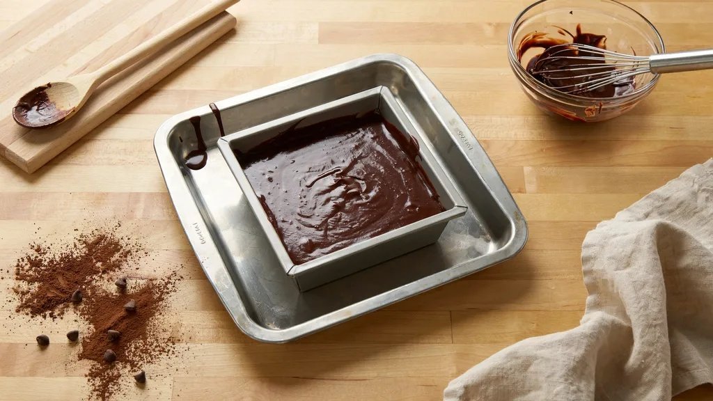 Chocolate brownie batter poured into a square metal baking pan with cocoa powder and chocolate chips on the counter.