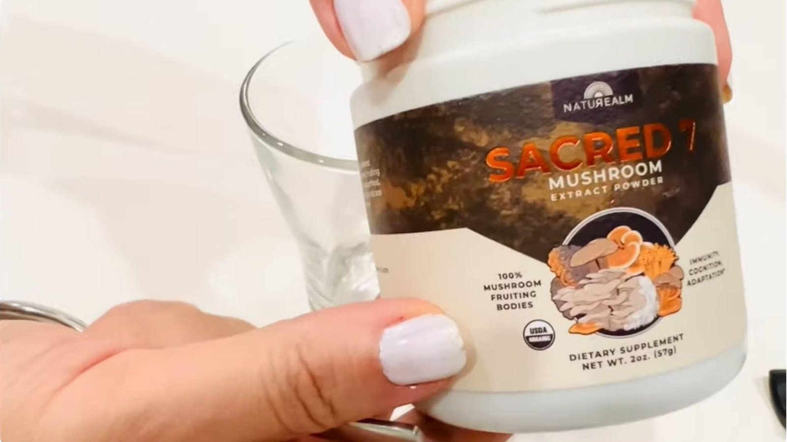 A hand holding a jar of Naturealm Sacred 7 Mushroom Extract Powder dietary supplement beside an empty clear glass mug.