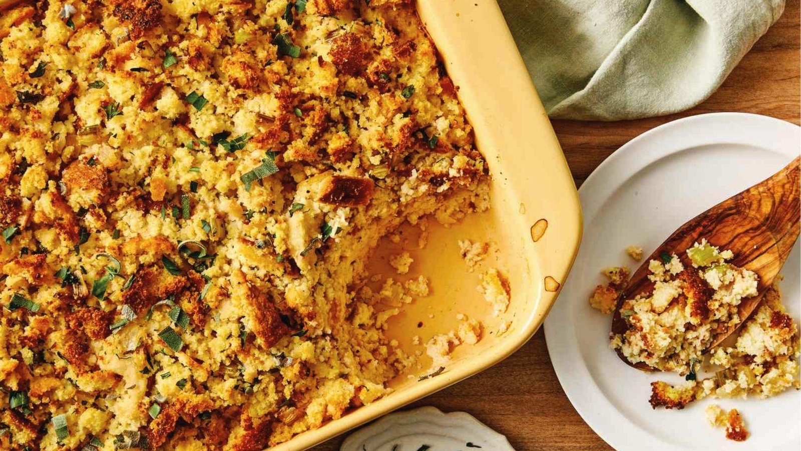 Scooped cornbread dressing with herbs in yellow casserole dish next to wooden spoon on plate