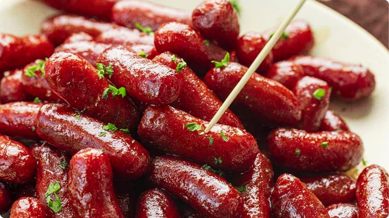 A close-up of glazed cocktail sausages piled high and garnished with fresh parsley, with one toothpick inserted.