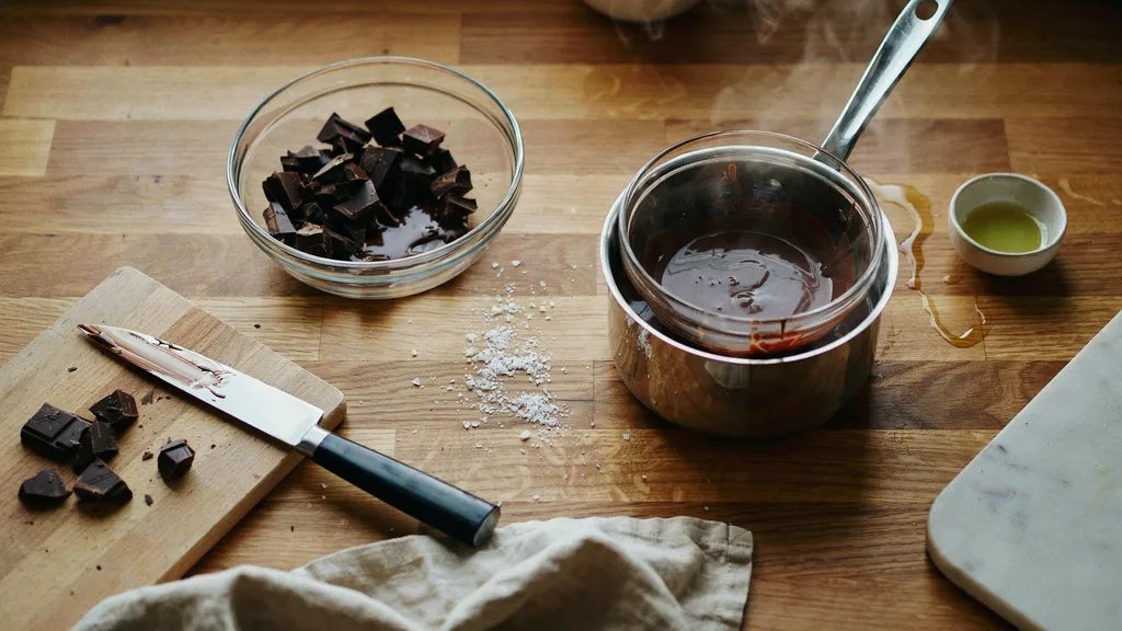 Dark chocolate chunks in a glass bowl beside a double boiler with melted chocolate on a wooden kitchen counter.