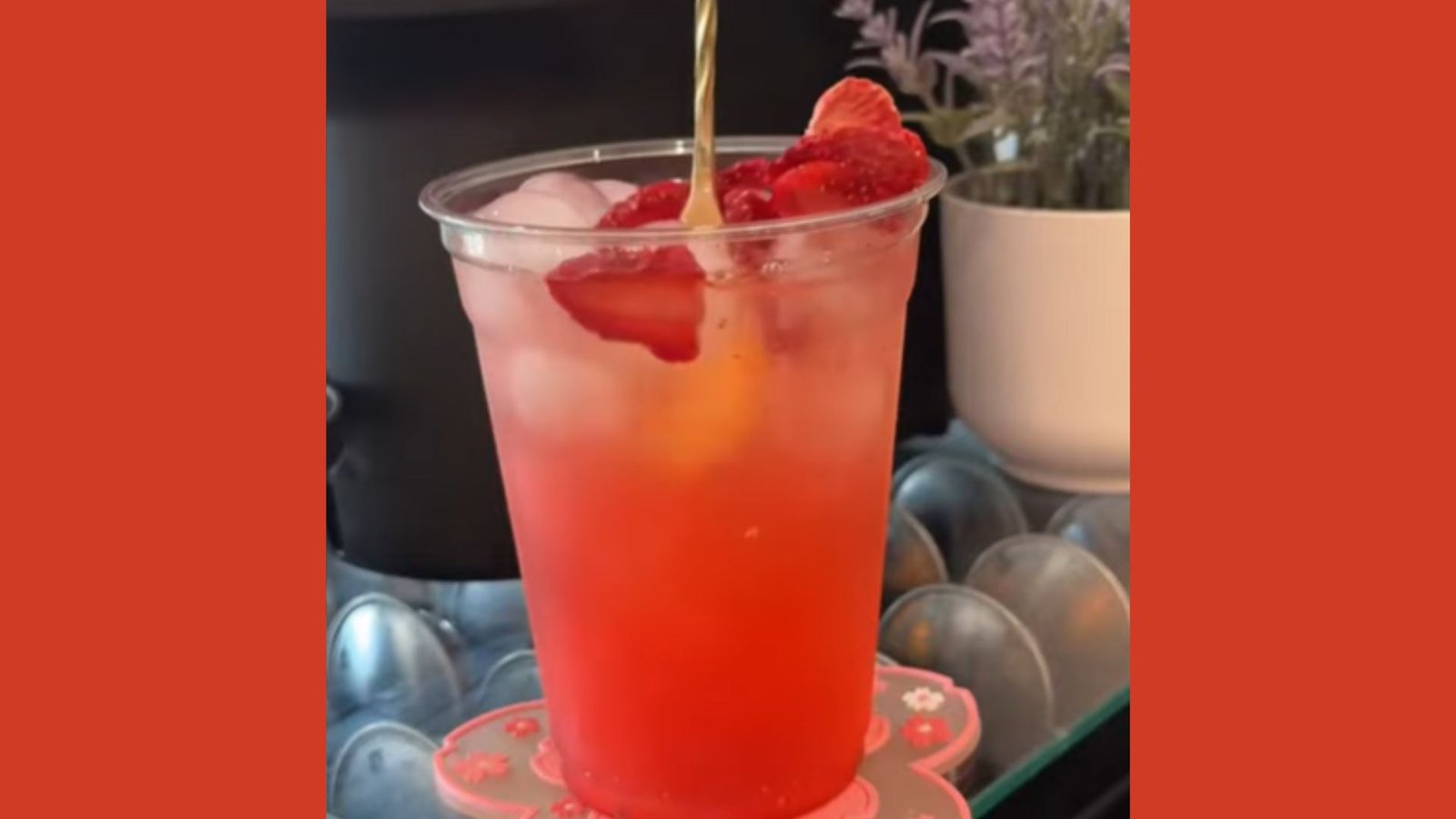  A clear plastic cup of layered pink-to-red strawberry iced drink topped with fresh strawberries and a gold straw mixing it.