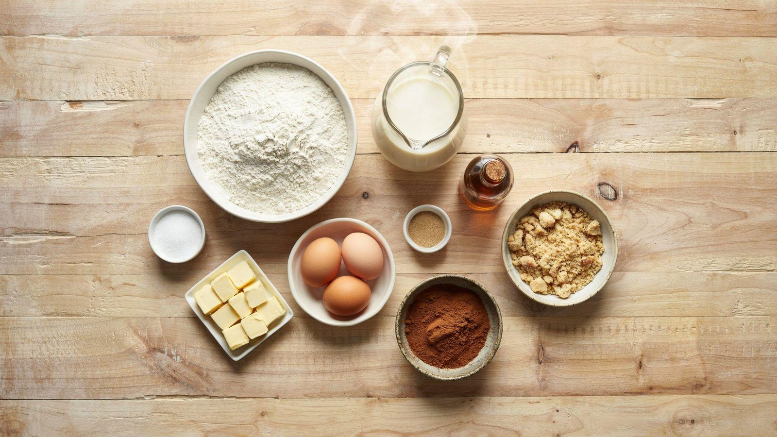 Flat lay of German coffee cake ingredients on a rustic wood surface: flour, eggs, butter, milk, cinnamon, and streusel crumble.