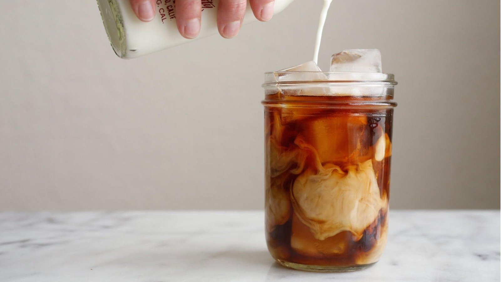 A hand pouring cream from a glass bottle into a mason jar of iced black coffee creating swirling patterns on marble.