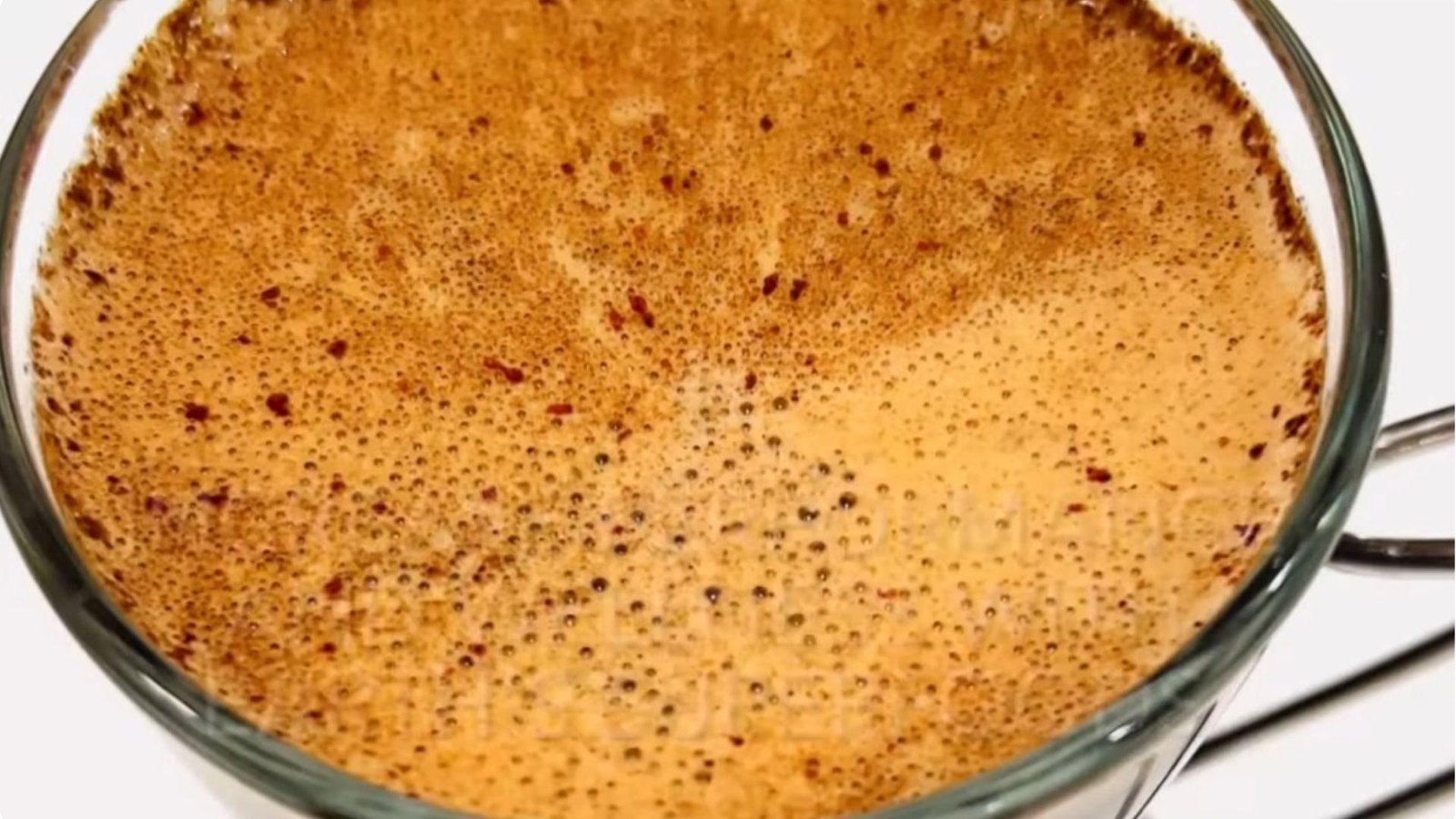 Overhead close-up of a frothy golden-brown mushroom coffee with fine bubbles in a clear glass mug on a white surface.