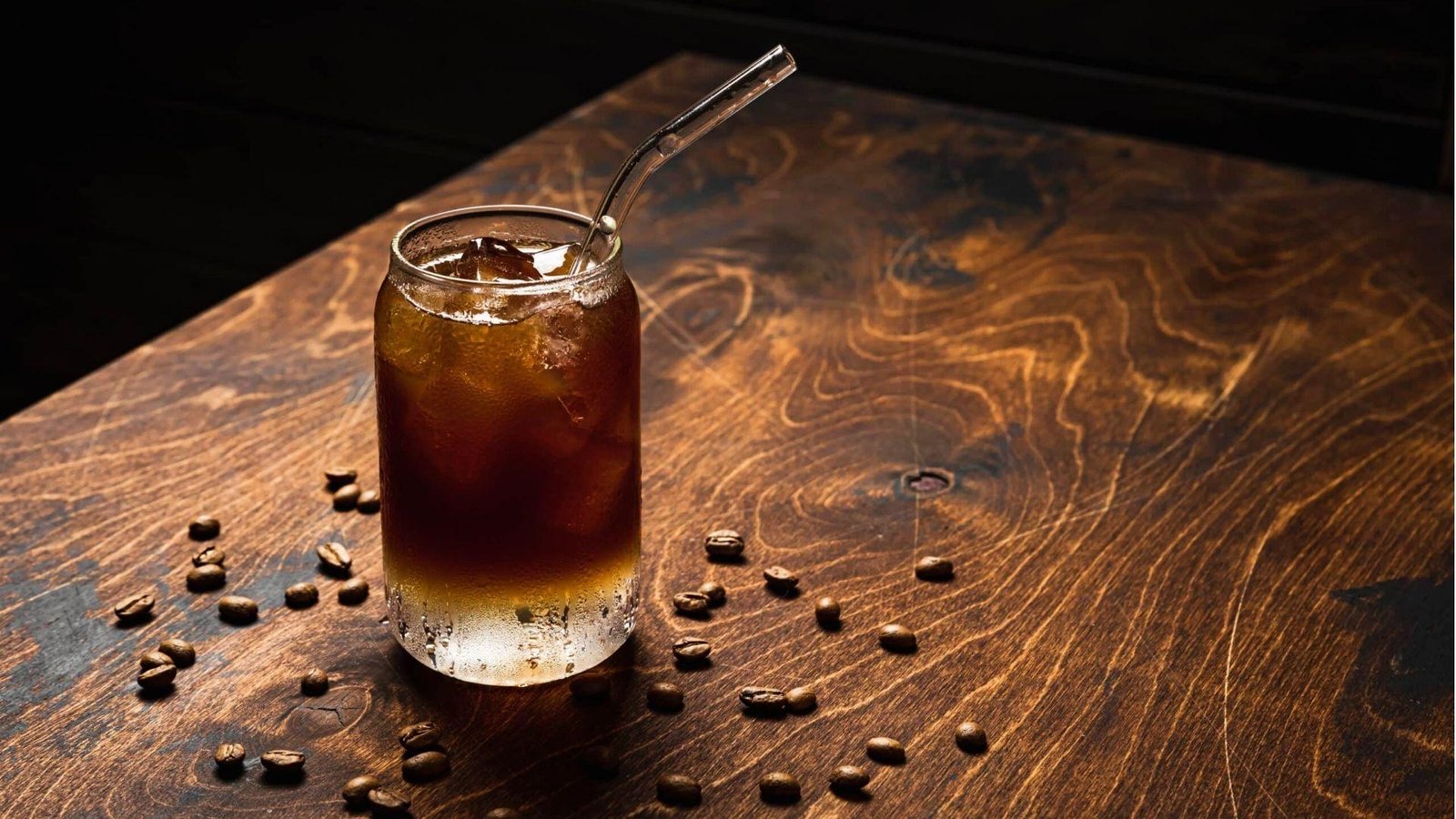 A can-shaped glass of cold brew coffee with ice and a metal straw, surrounded by coffee beans on a dark wooden surface.