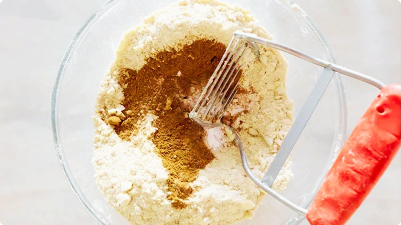Glass bowl with flour and brown cinnamon sugar being mixed with a pastry cutter with a red handle.