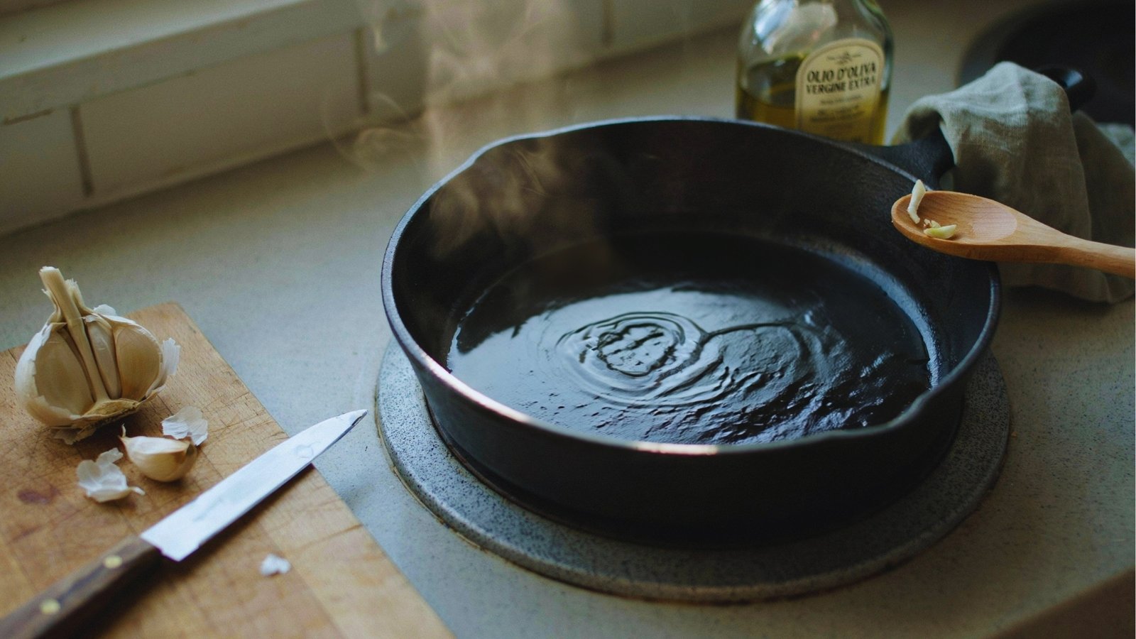 Steaming olive oil shimmers in black cast iron skillet beside a garlic bulb, knife, wooden spoon, and bottle on a sunlit stovetop.