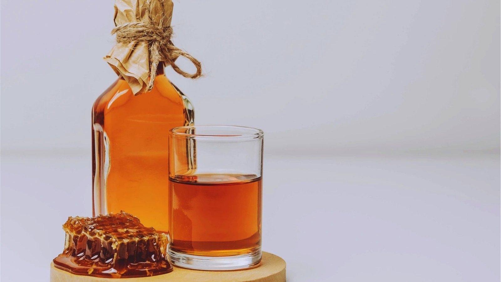  Amber honey mead in glass and rustic bottle sealed with paper and twine sits beside fresh honeycomb on wooden board