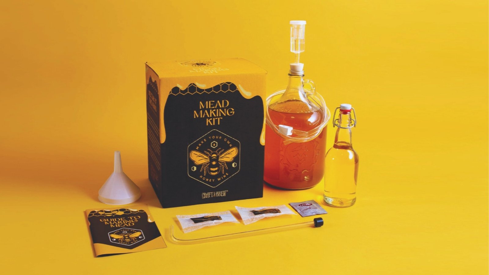 Black and yellow mead making kit with honey bee logo beside glass jug, swing-top bottle, funnel, airlock, and instructions on yellow background. 
