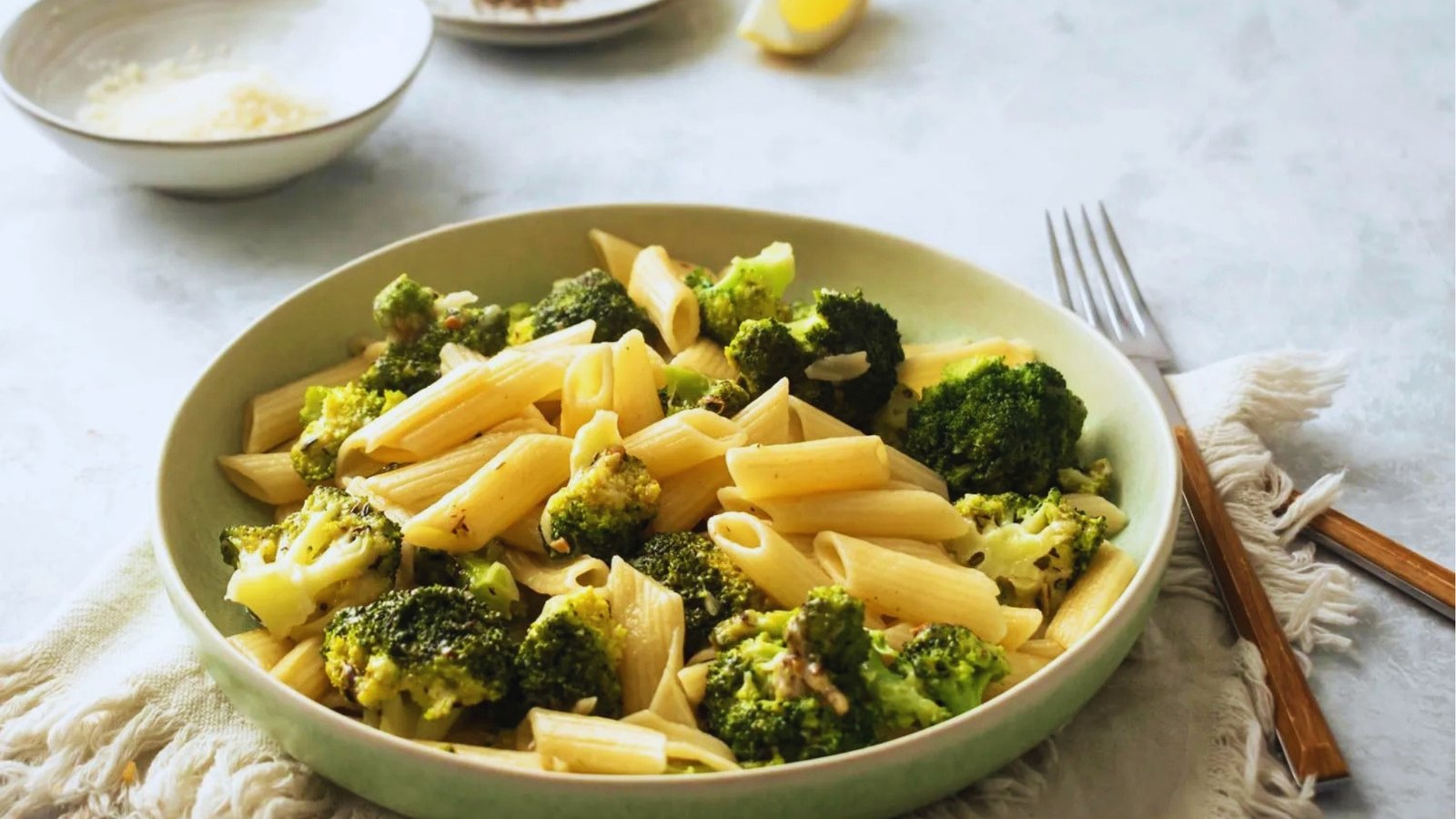 Green bowl of penne pasta tossed with roasted broccoli florets sits on linen cloth beside fork, lemon wedge, and parmesan.
